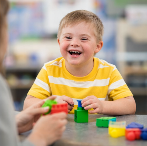 A young boy with Down syndrome playing with colorful blocks, smiling at an adult seated across the table in a classroom setting.