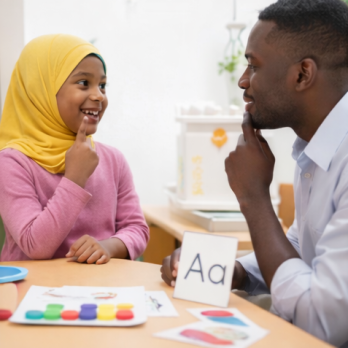 A man and a young girl wearing a hijab practicing speech sounds together, both smiling and pointing to their mouths at a table with learning cards.